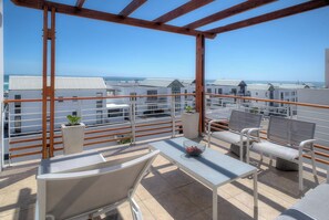 A rooftop terrace with a wooden pergola, seating area, and a view of the sea and buildings. - Charming 2-bedroom apartment in Big Bay near top spots (Cape Town)
