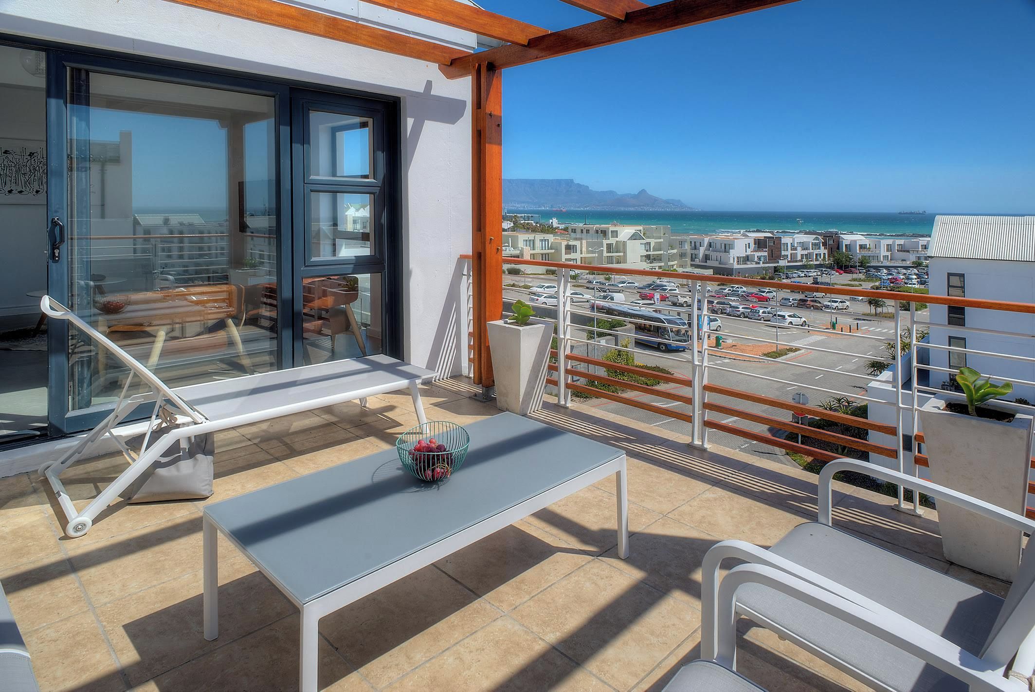 A balcony with a table, chairs, and a view of a coastal area with buildings and a parking lot.