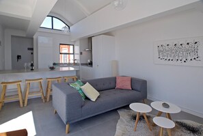 A modern living room with a grey sofa, wooden stools, and a kitchen in the background. - Charming 2-bedroom apartment in Big Bay near top spots (Cape Town)