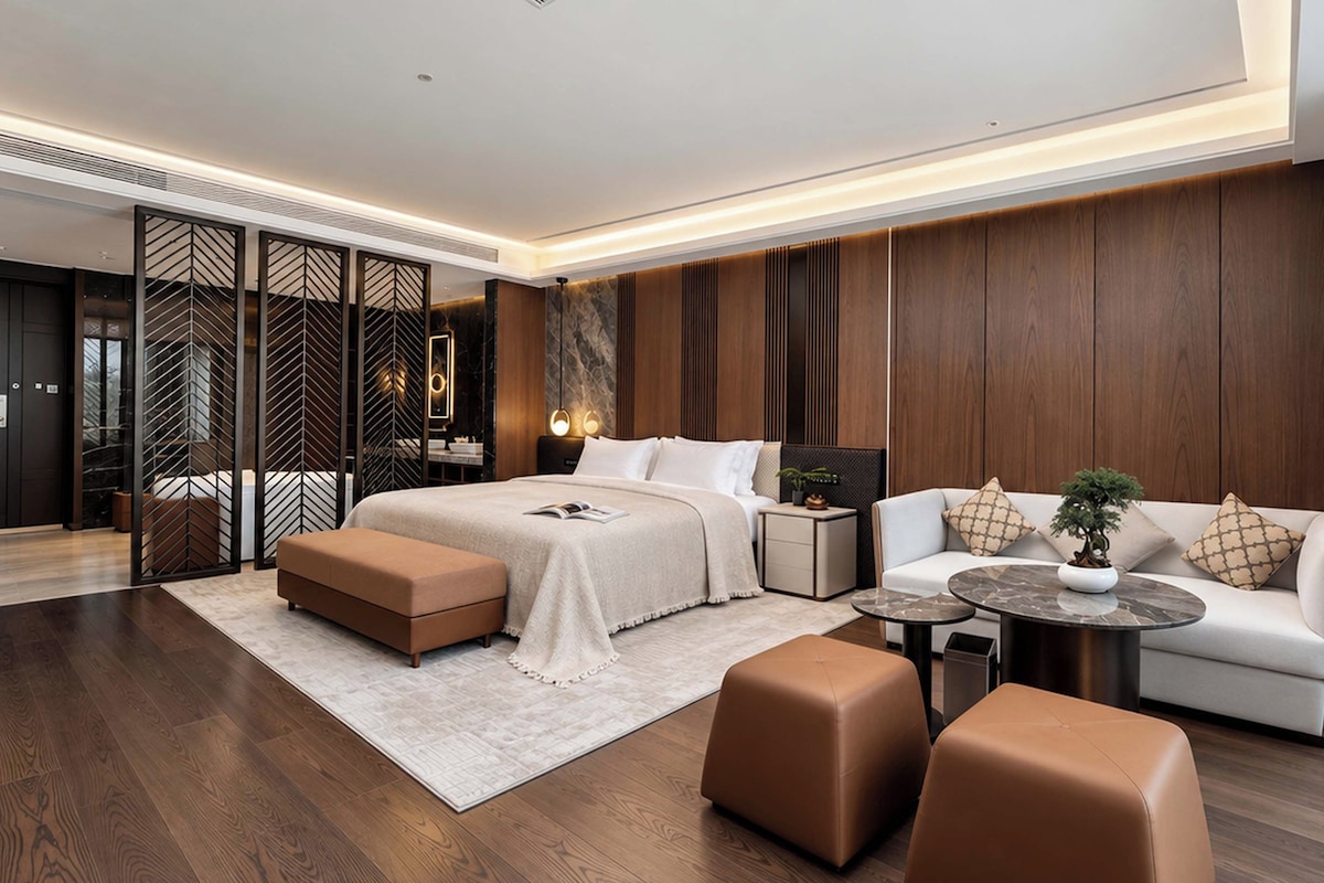 A modern hotel room with a large bed, a sofa, two ottomans, a small table, and a decorative plant.
