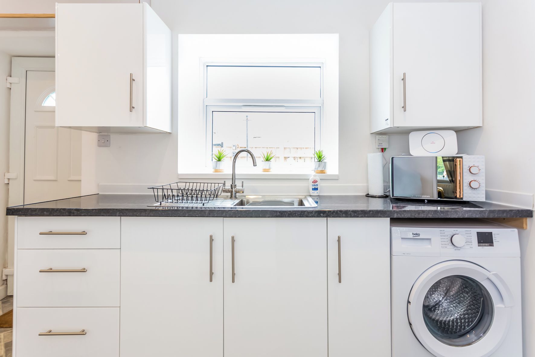A modern kitchen with a built-in washing machine, a sink with a dish rack, and a window with potted plants.