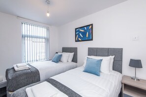 A hotel room with two beds, a window with blinds, a wall-mounted painting, a bedside lamp, and a small table. - Toast Stays - Greenwell (Darlington)