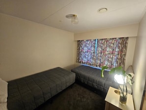 Ein Zimmer mit zwei Einzelbetten, einem Fenster mit blumigen Vorhängen, einem Nachttisch mit Lampe und einer Topfpflanze.