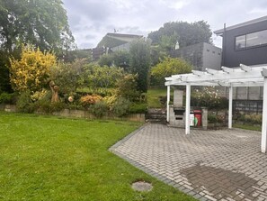 Ein Garten mit einer gepflasterten Terrasse, einer Pergola und verschiedenen Pflanzen und Bäumen.