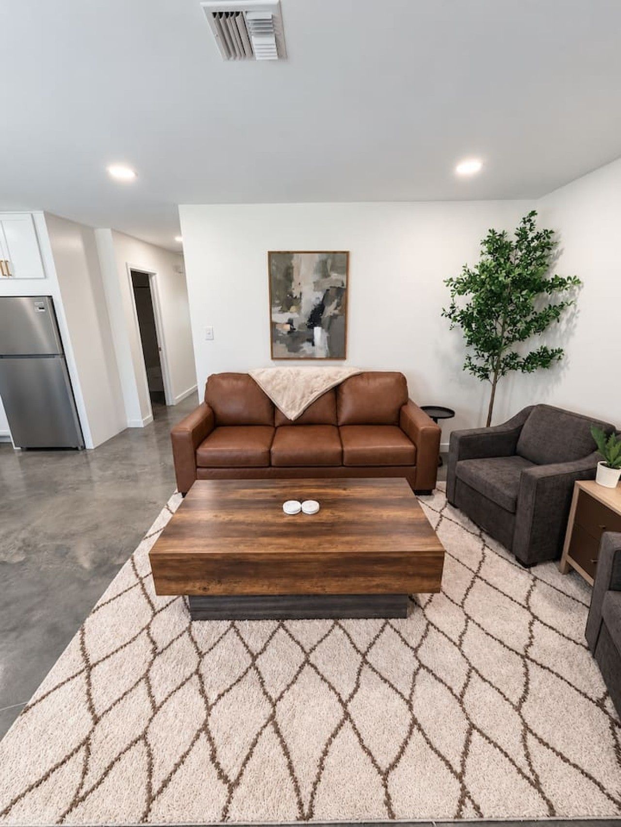 A living room with a brown leather sofa, a wooden coffee table, a patterned rug, a gray sofa, a potted plant, and a painting on the wall.