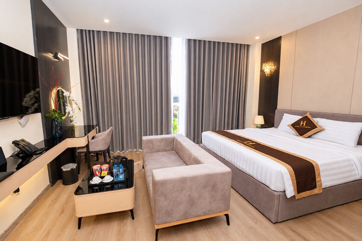 Habitación de hotel con cama, escritorio, silla, sofá, televisor y ventana con cortinas.