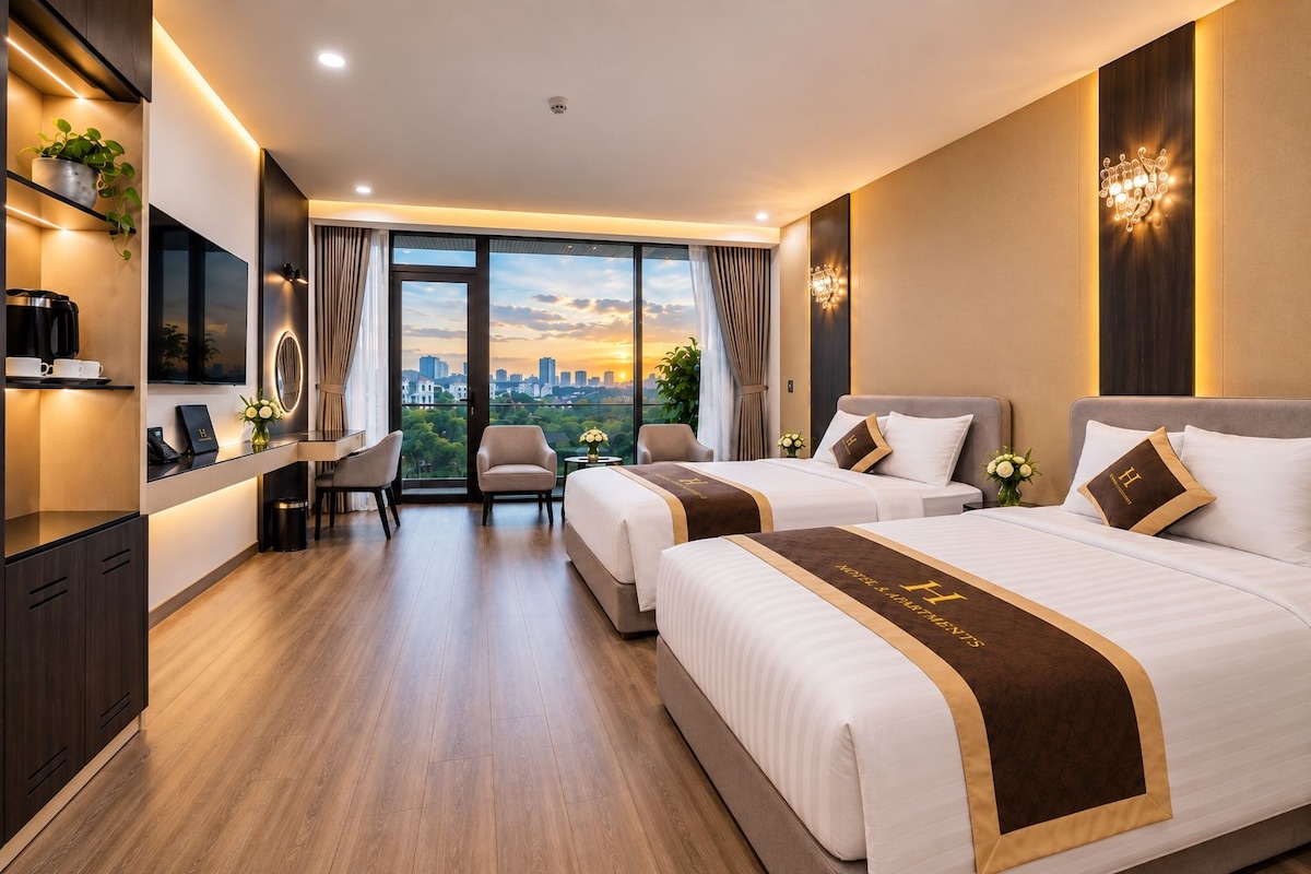 A hotel room with two beds, a desk, a chair, and a view of the cityscape.