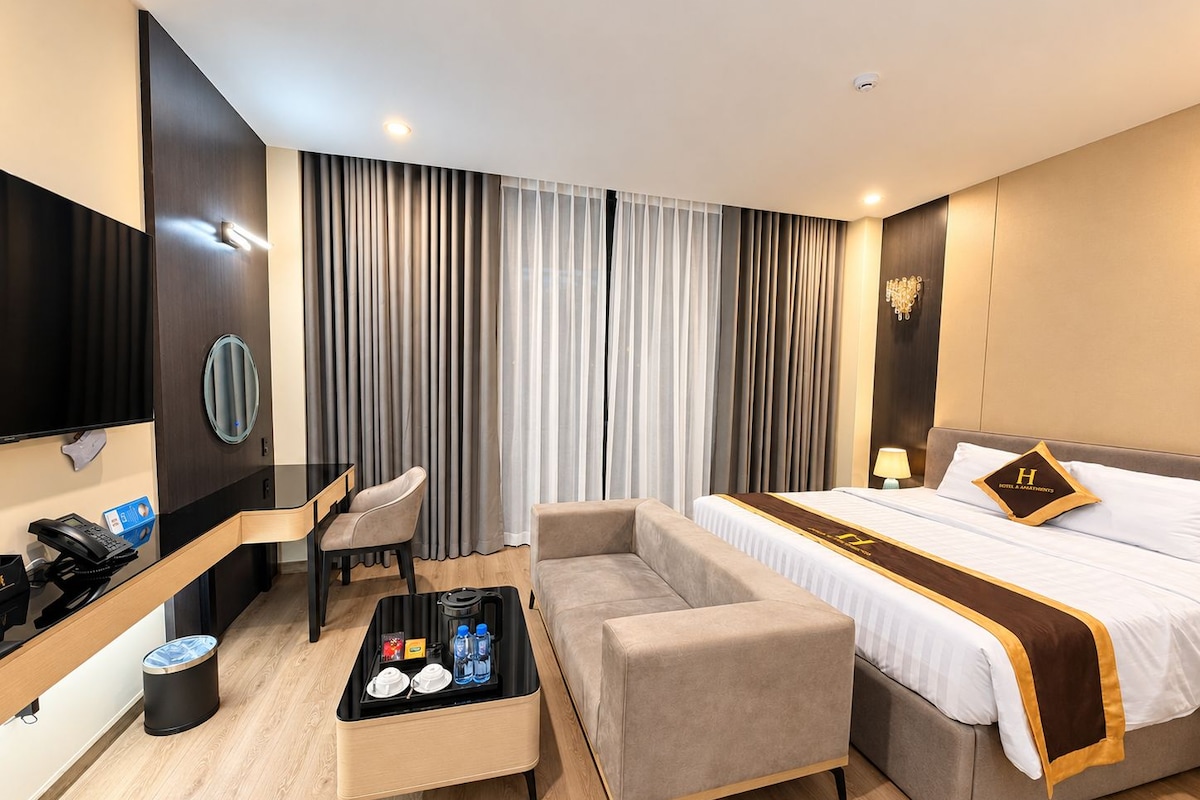 A hotel room with a bed, a desk, a chair, a sofa, and a small table with amenities.