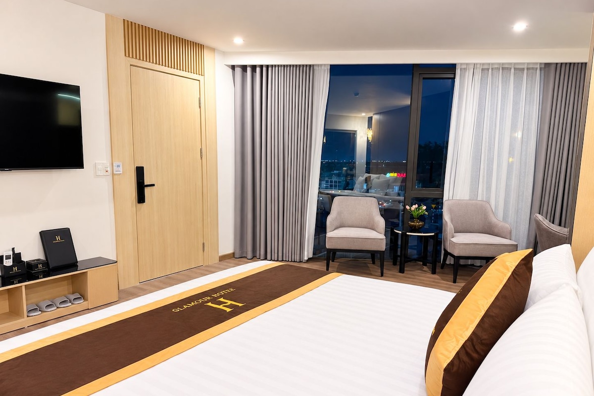 A hotel room with a large bed, a TV, a desk with a computer, and a view of the city through the window.