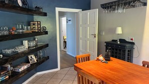 A dining area with a wooden table and chairs, a shelf with glassware, and a door leading to another room. - Sedgefield Lagoon & Sea Vista – 2 Bedroom Lower Garden Suite (Sedgefield)