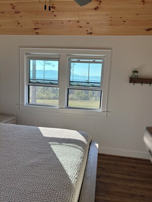 Quarto com cama grande, duas janelas, teto de madeira e uma pequena prateleira.
