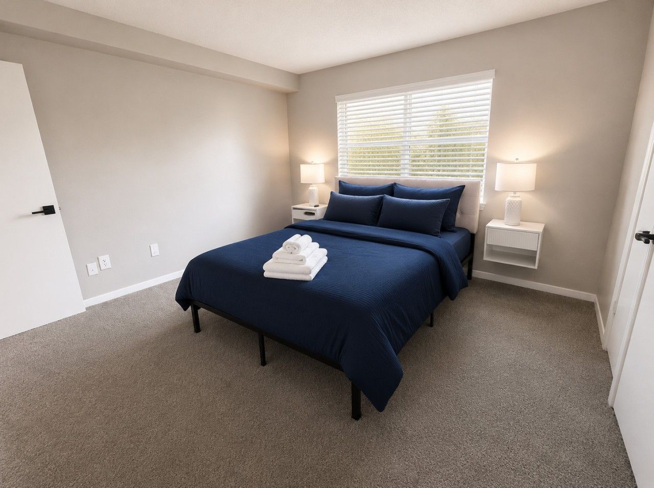 A bedroom with a bed, two bedside tables, a window with blinds, and a carpeted floor.