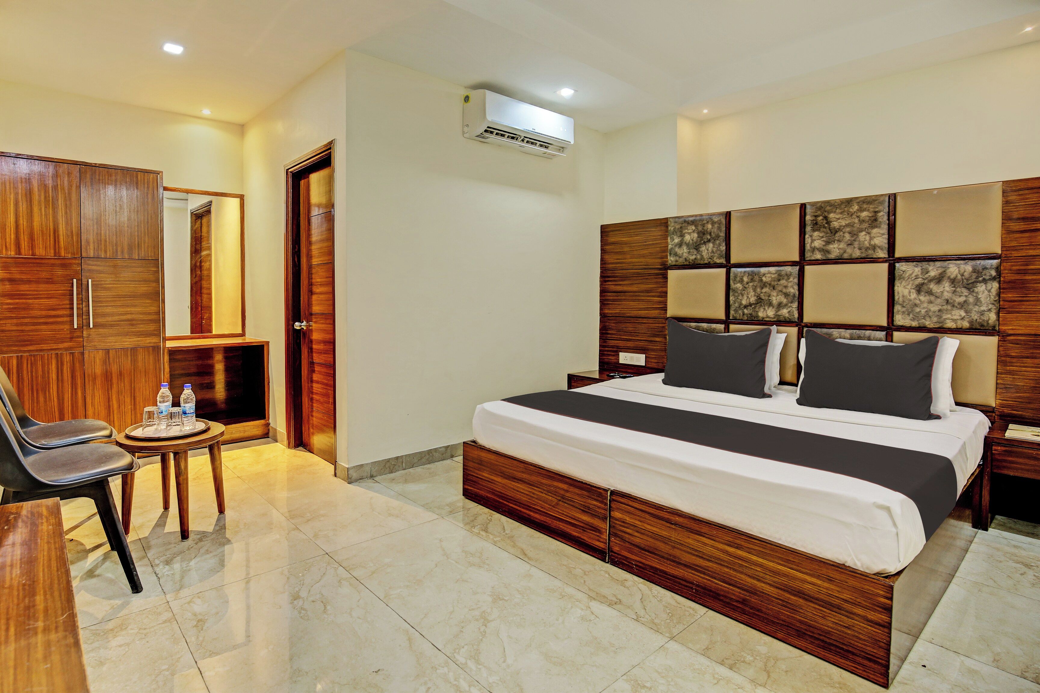 Una habitación de hotel con una cama grande, un armario de madera, una mesita con una silla y un equipo de aire acondicionado en la pared.