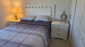 A bed with a striped comforter, two pillows, a bedside lamp, a white nightstand with drawers, and a wall clock. - Cozy 2-bedroom house in the villages. (Wildwood)