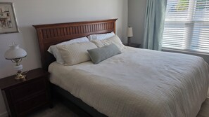 A neatly made bed with a wooden headboard, a bedside table with a lamp, and a window with blinds. - Cozy 2-bedroom house in the villages. (Wildwood)