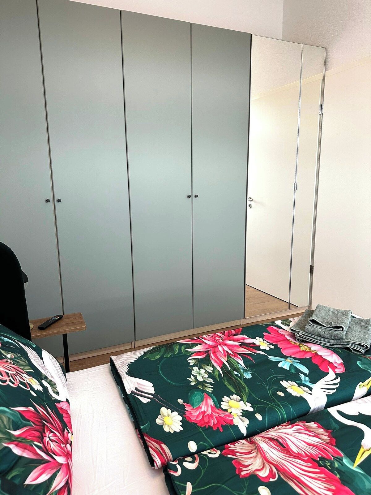 A bedroom with a grey wardrobe, a wooden bedside table, and a bed with floral bedding.