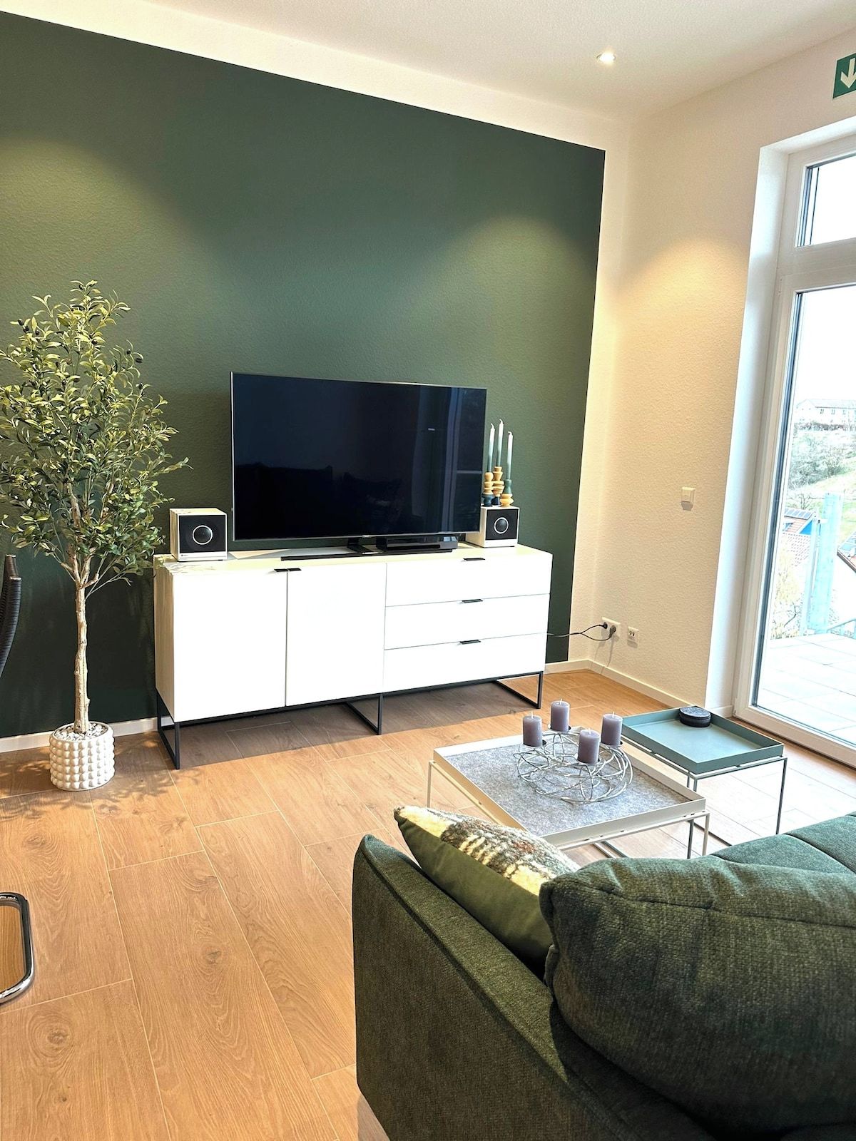 A modern living room with a flat-screen TV, a white cabinet, a green sofa, and a potted plant.