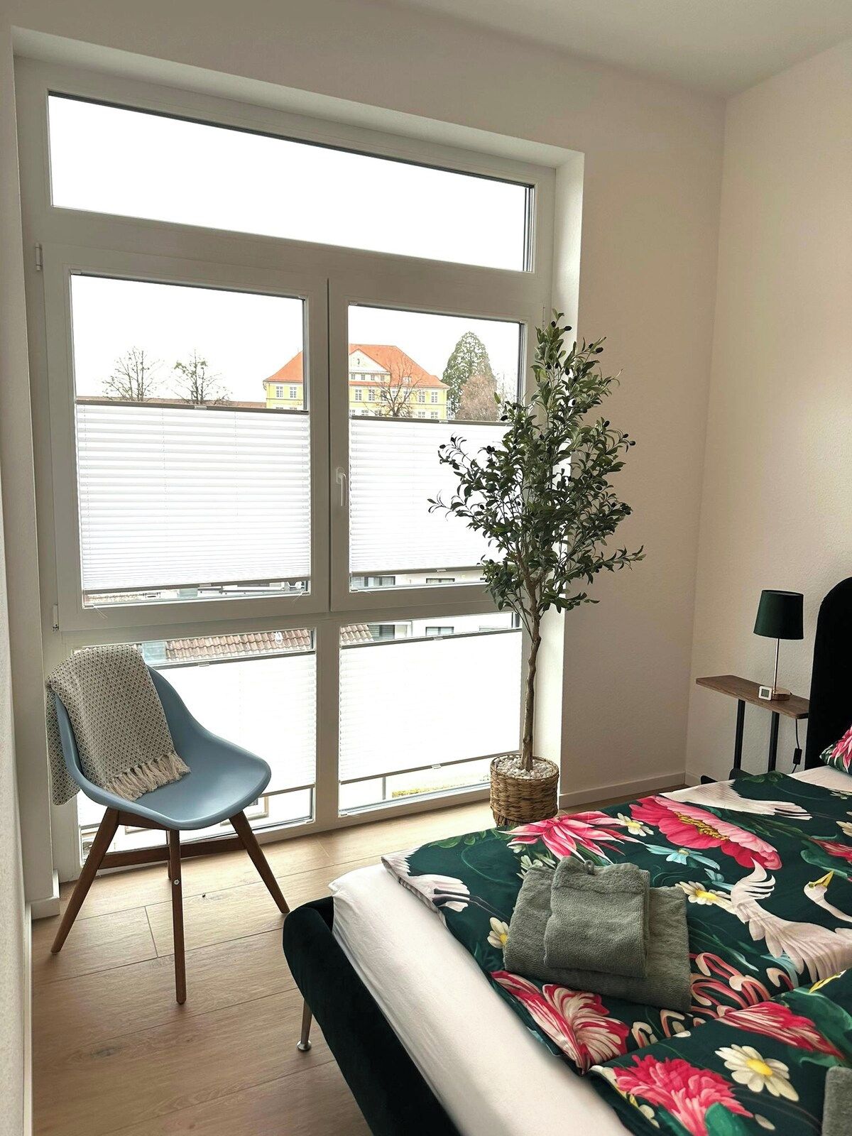 Ein Schlafzimmer mit großem Fenster, einem Sessel, einem Bett mit Blumenmuster auf der Bettwäsche, einer Topfpflanze und einem Nachttisch mit Lampe.