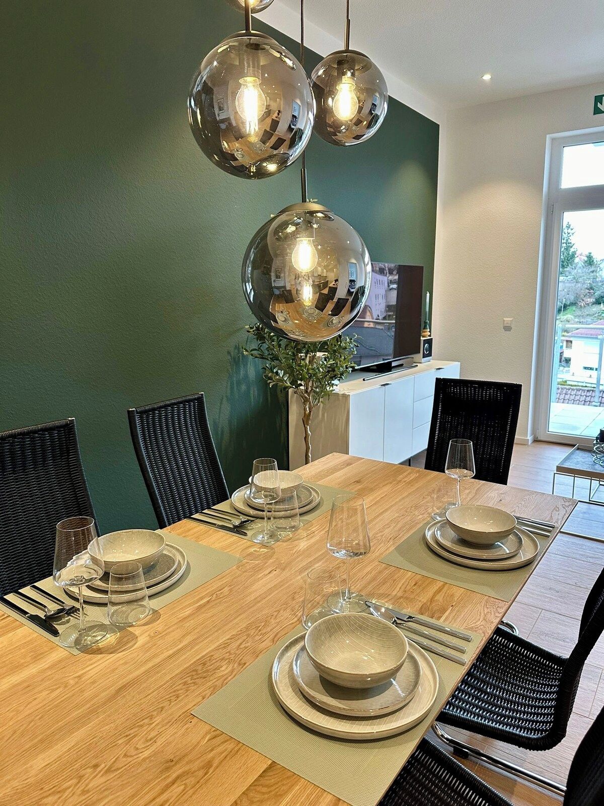 A dining area with a wooden table set for four, surrounded by black chairs, and a green wall in the background.