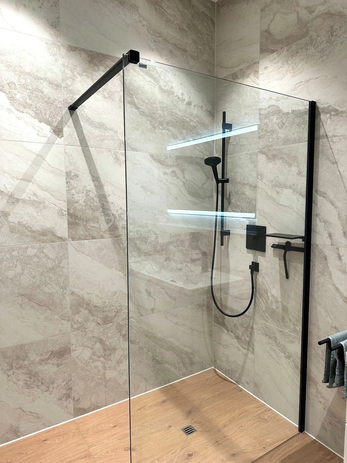Eine Dusche mit Glasabtrennung, Holzboden, einer Handbrause und einer an der Wand montierten Bedienkonsole.
