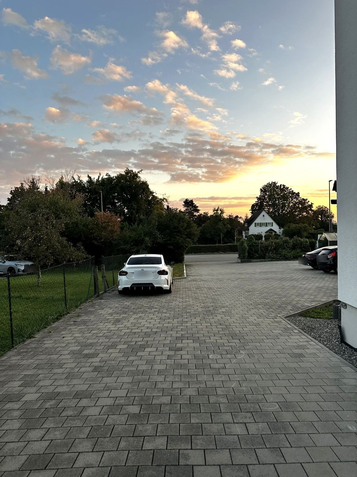 Une allée pavée mène à un bâtiment blanc, entouré d’une clôture et bordé de voitures garées.