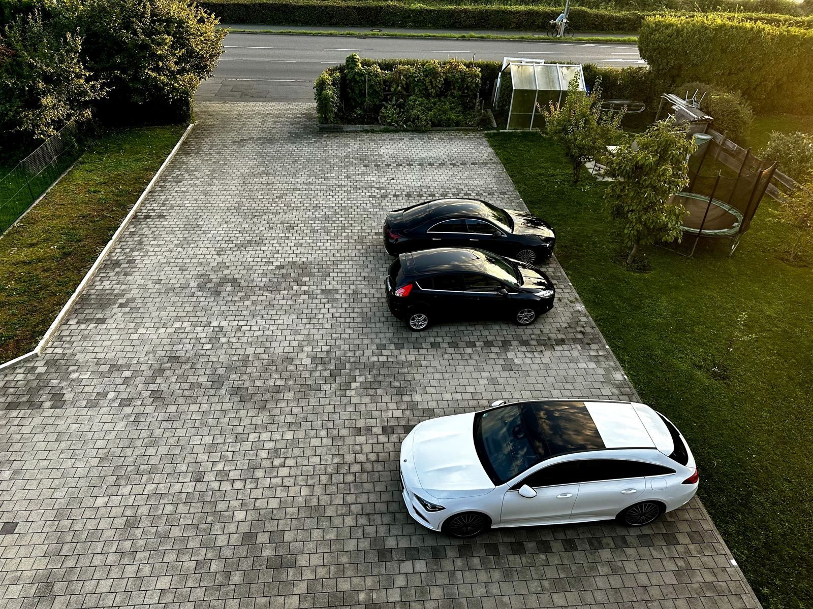A paved driveway with three parked cars, a grassy area, and a hedge.
