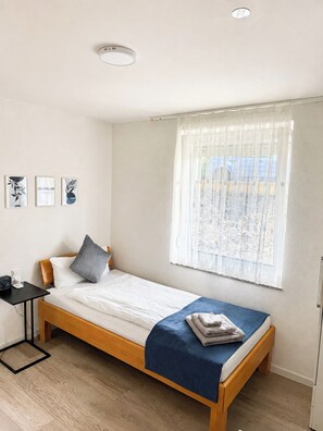 A single bed with a blue blanket, a bedside table with a fan, and a window with sheer curtains. - STAY | Ruhig für 1 · 300 m zum See · Parkplatz (Friedrichshafen)