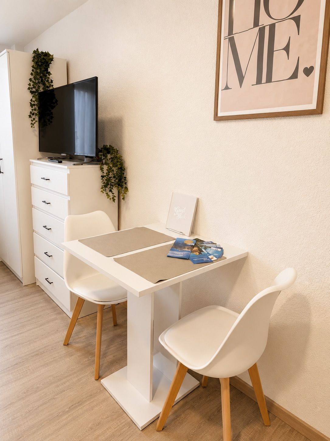 Ein schlichtes Zimmer mit einem weißen Schreibtisch, zwei weißen Stühlen, einem Flachbildfernseher auf einem Schrank und einem gerahmten Motivationszitat an der Wand.