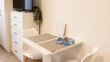 Ein schlichtes Zimmer mit einem weißen Schreibtisch, zwei weißen Stühlen, einem Flachbildfernseher auf einem Schrank und einem gerahmten Motivationszitat an der Wand.