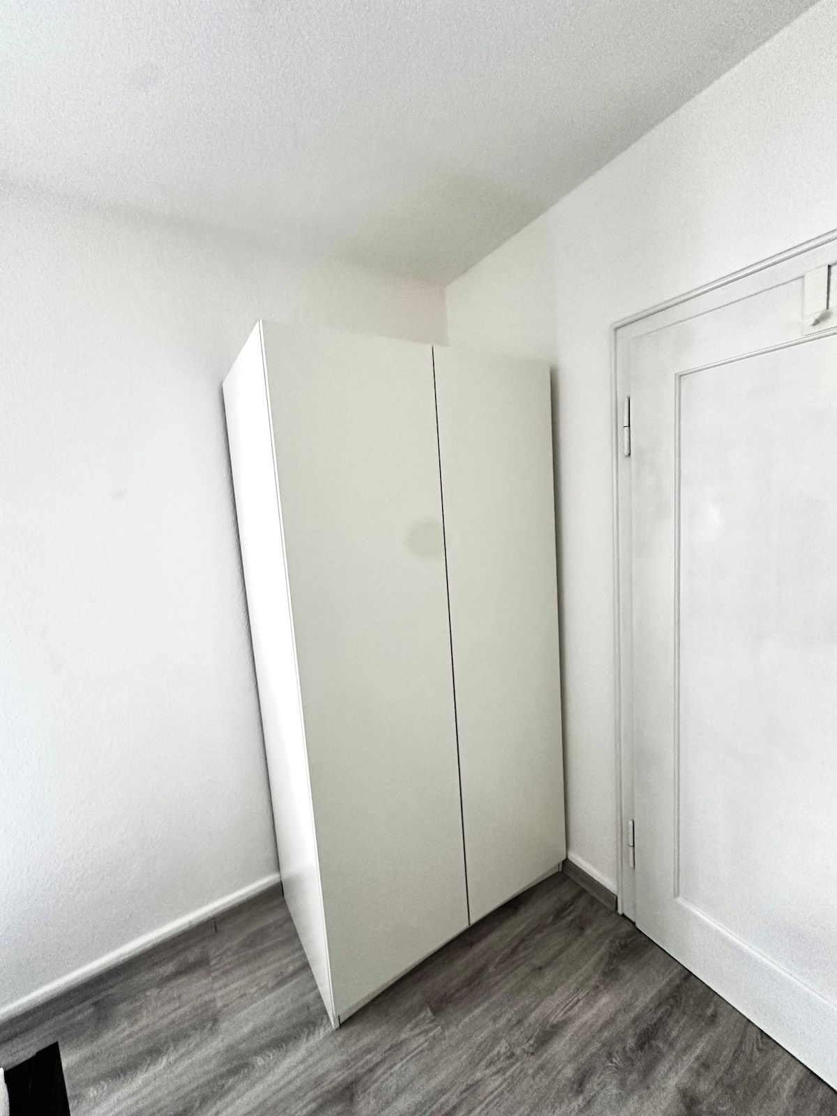 A room with a white wardrobe, a door, and a grey wooden floor.