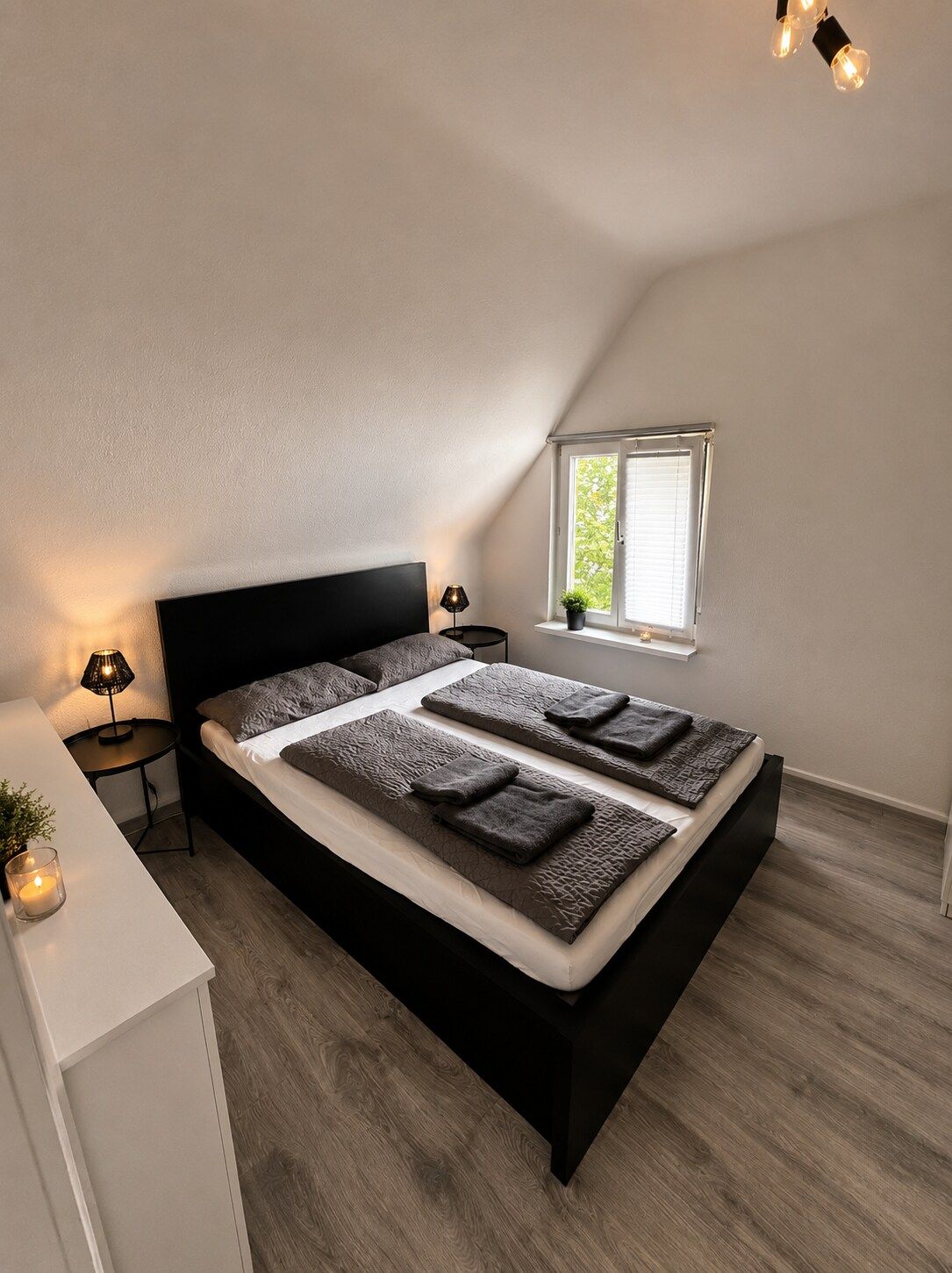 Ein Schlafzimmer mit einem schwarzen Bett, einem Nachttisch mit Lampe, einem Fenster mit Jalousien und einer kleinen Pflanze auf der Fensterbank.