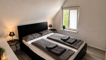 Ein Schlafzimmer mit einem schwarzen Bett, einem Nachttisch mit Lampe, einem Fenster mit Jalousien und einer kleinen Pflanze auf der Fensterbank.