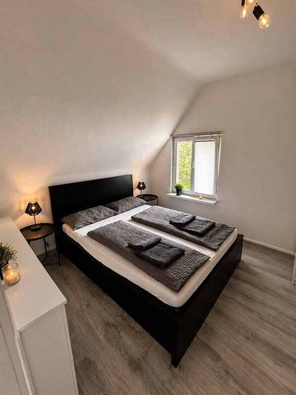Ein Schlafzimmer mit einem schwarzen Bett, einem Nachttisch mit Lampe, einem Fenster mit Jalousien und einer kleinen Pflanze auf der Fensterbank.