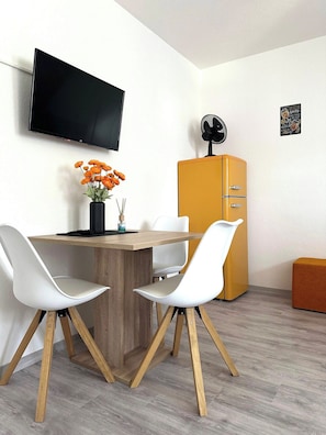 Ein modernes Zimmer mit einem Holzespeisetisch, zwei weißen Stühlen, einem an der Wand befestigten Fernseher, einem Retro-Kühlschrank und einem kleinen Ventilator.