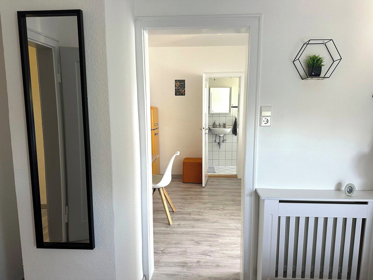 Ein Flur, der zu einem Badezimmer mit einem weißen Heizkörper, einem weißen Stuhl und einer sechseckigen Wanddekoration führt.