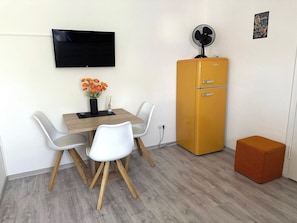 Un espacio habitable compacto con un televisor de pantalla plana montado en la pared, una pequeña mesa con sillas, un refrigerador de estilo vintage y un pouf naranja.