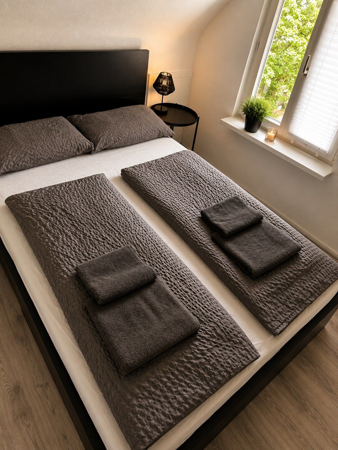 A modern bedroom with a large bed, a bedside table, a lamp, and a window with a view of greenery.
