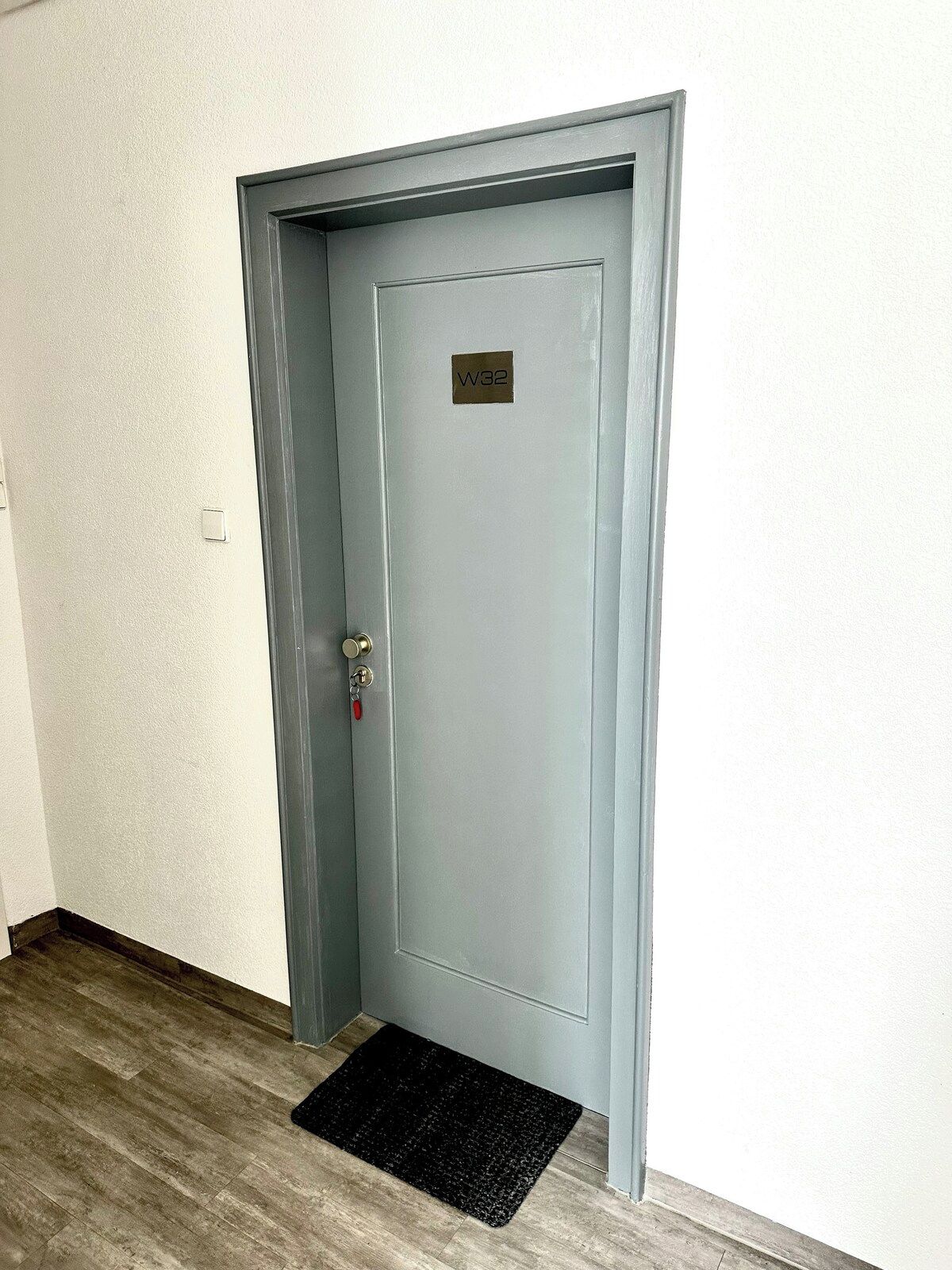 Una puerta cerrada con un ojo de cerradura y un timbre, un felpudo y un interruptor de luz en la pared.