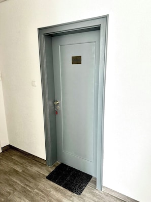 A closed door with a keyhole and a doorbell, a doormat, and a light switch on the wall. - STAY | City-Apartment W32, Friedrichshafen (Friedrichshafen)