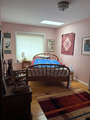 Ein Schlafzimmer mit einem hölzernen Bett, einem Fenster mit Jalousien, einem Nachttisch mit Lampe und einem Wandregal mit Dekorationen.