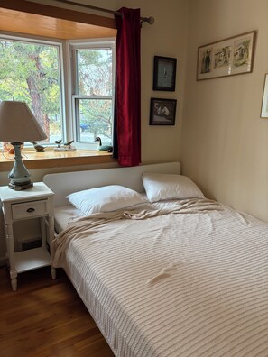 Ein Schlafzimmer mit einem Bett, einem Nachttisch, einer Lampe, einem Fenster mit Blick auf Bäume und zwei gerahmten Bildern an der Wand.