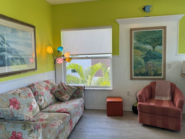 A room with a floral sofa, a pink armchair, a window with a view of palm trees, a painting, and a colorful floor lamp. - Old-Florida Resort, Steps to Beach & Village, Heated Pool (Siesta Key)