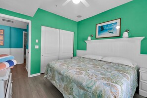 A bedroom with a bed, a white door, a painting, and a bathroom with a towel. - Sand & Sunsets Steps Away, Heated Pool, Village (Siesta Key)