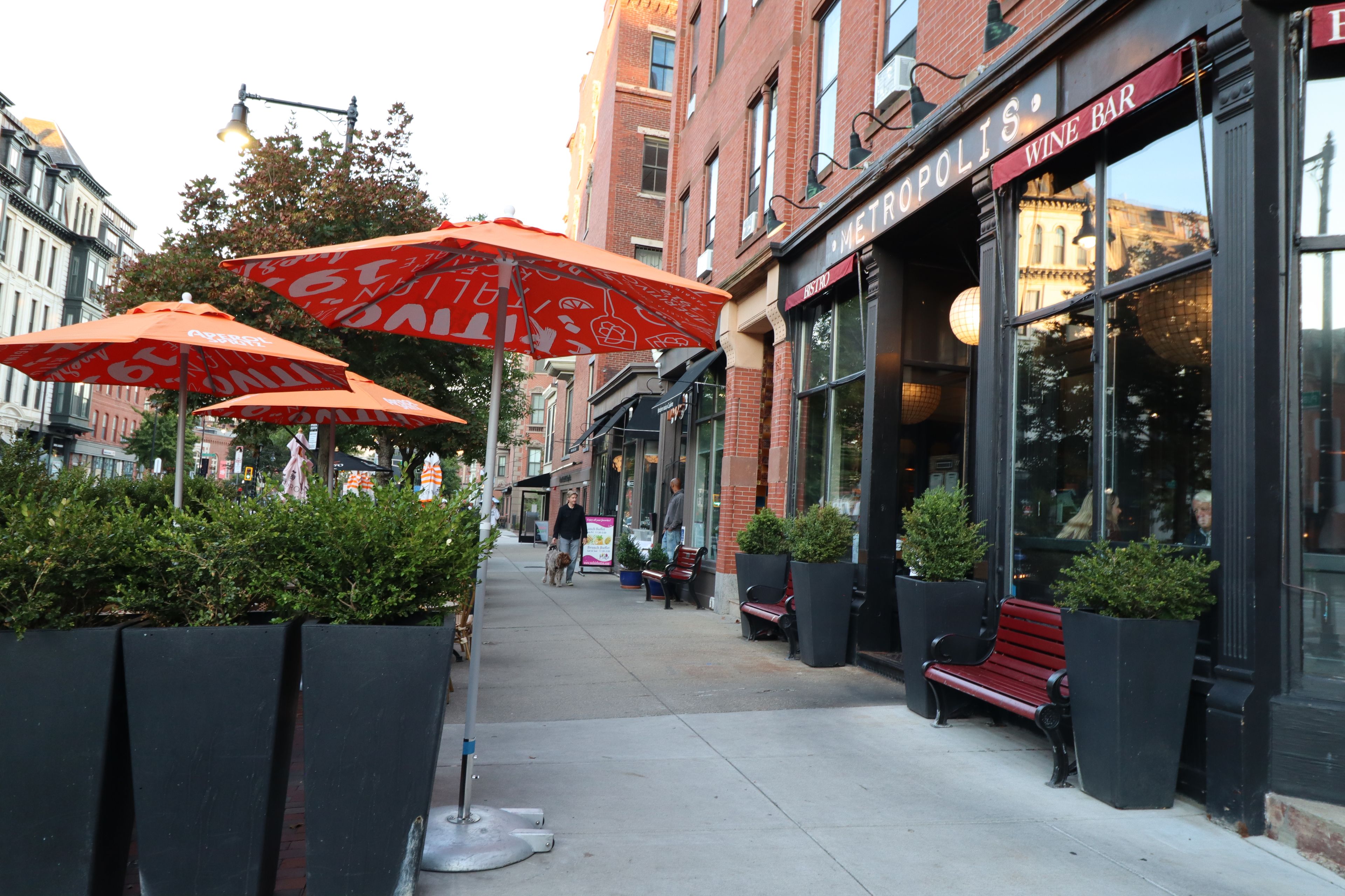 A sidewalk with outdoor seating and umbrellas, a storefront with a wine bar sign, and a brick building in the background.