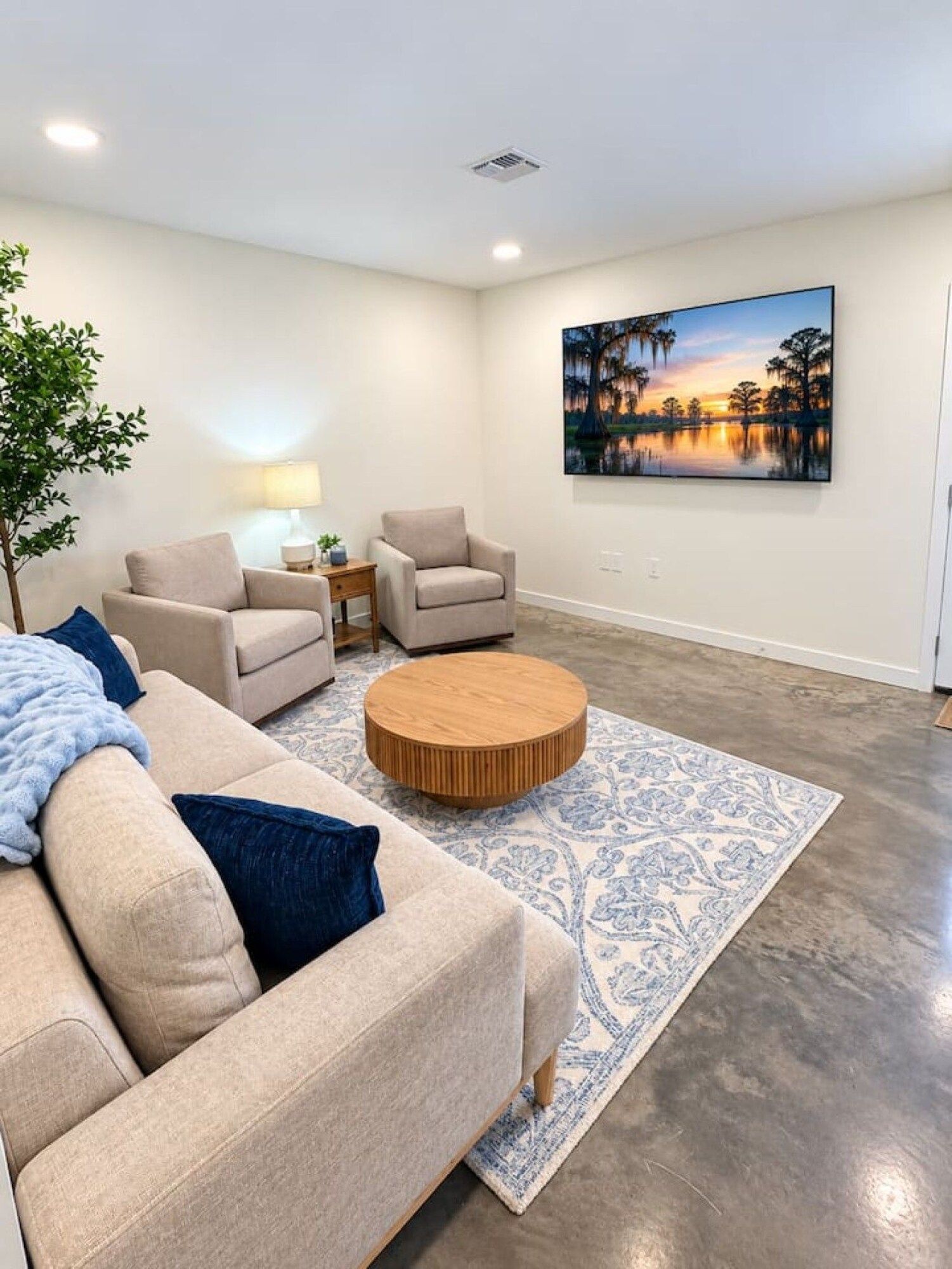 A modern living room with a beige sofa, two armchairs, a coffee table, and a large painting of a sunset over water.