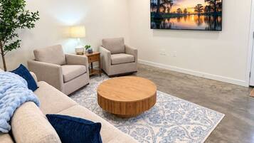 A modern living room with a beige sofa, two armchairs, a coffee table, and a large painting of a sunset over water.