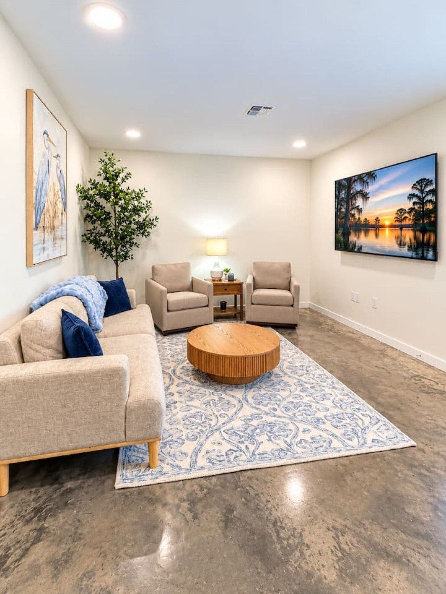 A living room with a beige sofa, two armchairs, a coffee table, a patterned rug, a painting, and a tree.