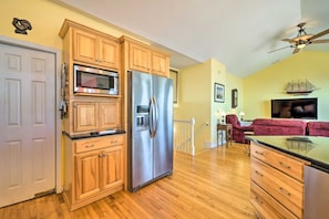 A kitchen with wooden cabinets, stainless steel appliances, a wooden floor, a dining area with a red sofa, and a ceiling fan. - Waterfront Sneads Ferry Retreat w Deck & Bar! (Sneads Ferry)
