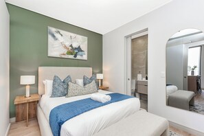 A modern bedroom with a large bed, a nightstand, a bedside table, a bathroom with a mirror, and a view of the outside. - One Rosebank | Contemporary Comfort (Johannesburg)