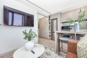 A modern living room with a TV, a white vase with green plants, a remote control, and a patterned rug. - One Rosebank | Contemporary Comfort (Johannesburg)